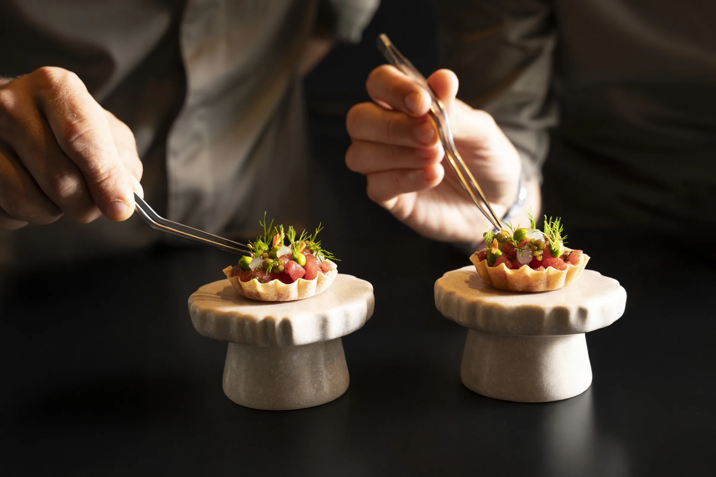 Chef's hands finishing tuna tartelettes with tweezers on stone pedestals